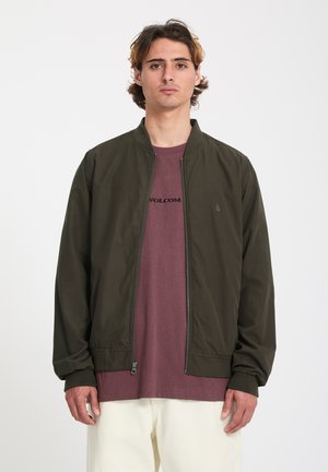 Jeune homme aux cheveux bruns mi-longs portant une veste bomber olive sur un t-shirt Volcom violet et un pantalon écru, debout devant un fond neutre.