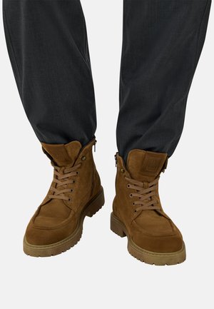 Botas de ante marrón con cordones, paneles texturizados, suelas de goma y cremalleras laterales, combinadas con pantalones gris oscuro.