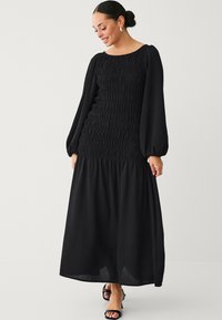 Schwarzes langärmliges Maxikleid mit gerafftem Oberteil und fließendem Rock. Glatter Stoff, runder Ausschnitt und luftige Ärmel. Getragen mit schwarzen Sandalen.