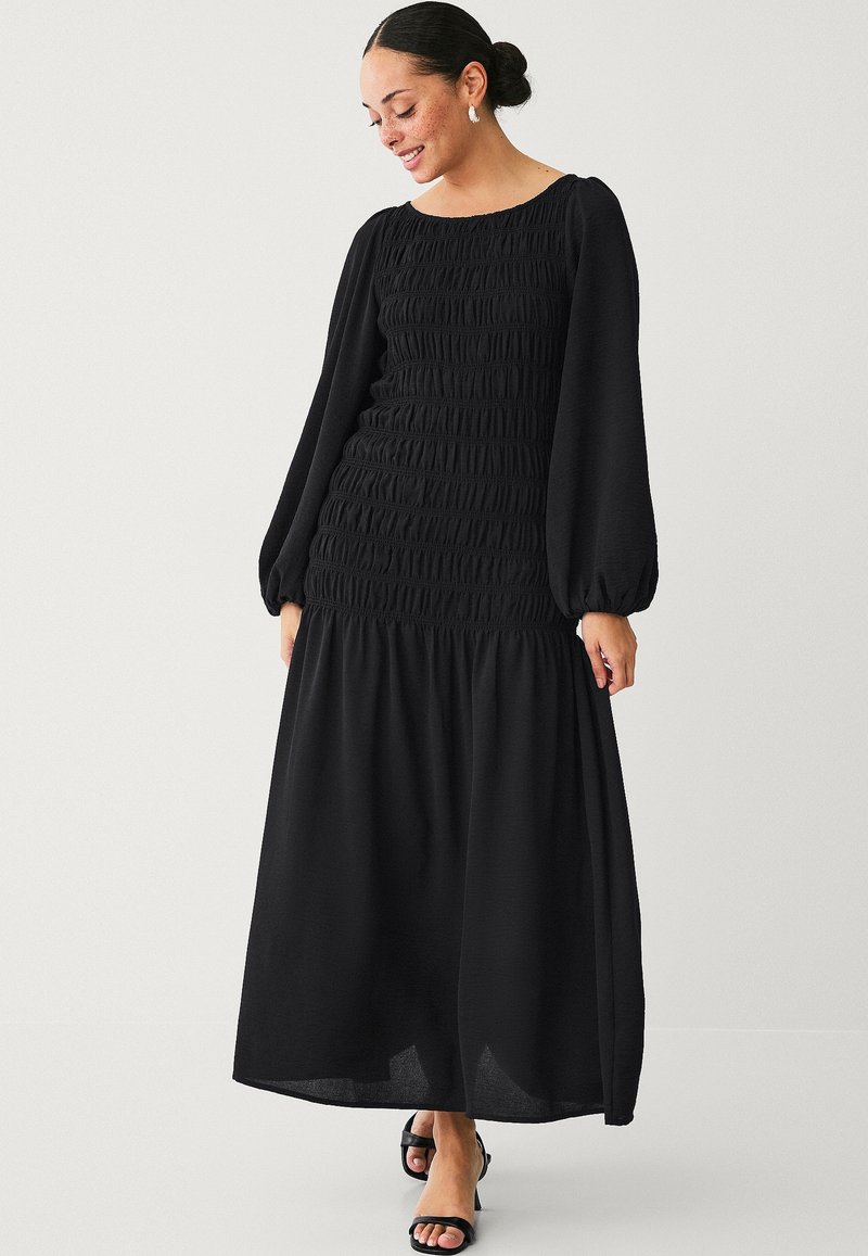 Schwarzes langärmliges Maxikleid mit gerafftem Oberteil und fließendem Rock. Glatter Stoff, runder Ausschnitt und luftige Ärmel. Getragen mit schwarzen Sandalen.