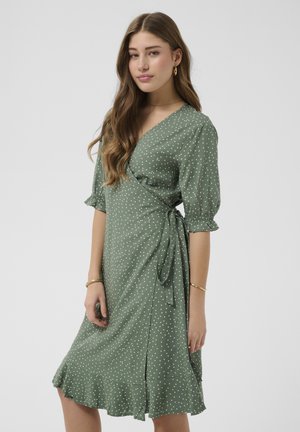 Młoda kobieta o długich falujących włosach, ubrana w zieloną sukienkę kopertową w groszki z falbaną na dole i rękawami do łokcia na jednolitym tle.