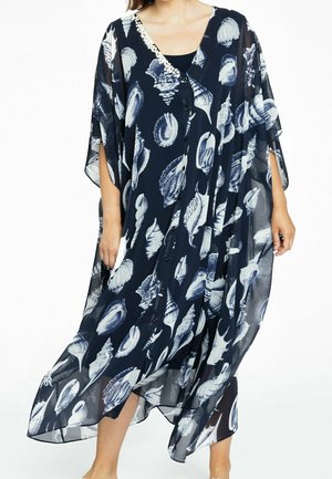 Femme portant une robe fluide bleu marine avec des imprimés de coquillages blancs et des boutons sur le devant, debout devant un fond blanc.