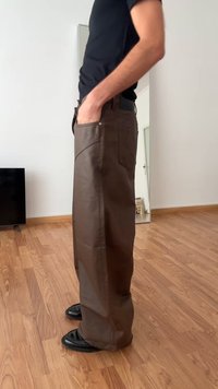 Hnedé kožené nohavice s rovnými nohavicami, obsahujúce bočné vrecká a hladký povrch. Nosia sa s čiernymi topánkami, stojac na drevenej podlahe.