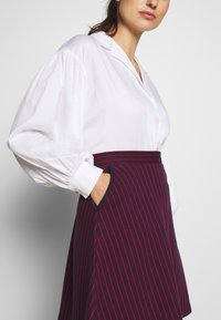 Camicia bianca con maniche a palloncino abbinata a una gonna blu navy di lunghezza al ginocchio con righe rosse verticali e tasche laterali.