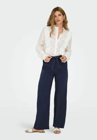 Blouse blanche brodée avec des détails à volants, associée à un pantalon large bleu marine. Sandales plates beige complètent la tenue. Design simple et élégant.