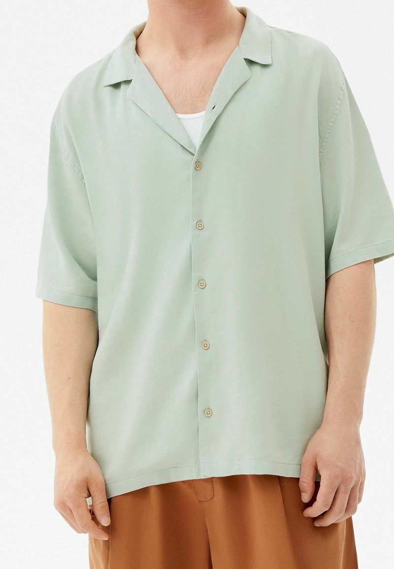 Homme portant une chemise verte claire à manches courtes boutonnée sur un débardeur blanc et un pantalon marron, debout devant un fond blanc.