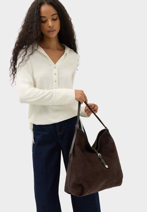 Femme aux longs cheveux bouclés portant un haut blanc boutonné et un pantalon foncé, tenant un grand sac à main marron foncé avec des anses d'épaule.
