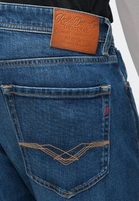 Sötétkék farmernadrág bőrből készült pánttal a derékrészen. Narancssárga varrással és díszítő mintával a hátsó zsebén.