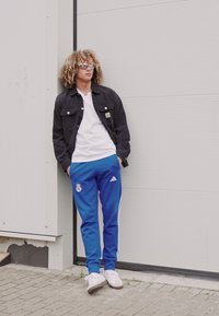 Black denim jacket, white t-shirt, blue athletic pants with white stripes and logo, and white sneakers. Outdoors against a neutral wall.