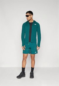 Άνδρας που φοράει τζάκετ με φερμουάρ σε τιρκουάζ και σορτς, μαύρη μπλούζα, μαύρες κάλτσες, μαύρα παπούτσια και μεγάλα σκούρα γυαλιά ηλίου, στέκεται μπροστά σε λευκό φόντο.