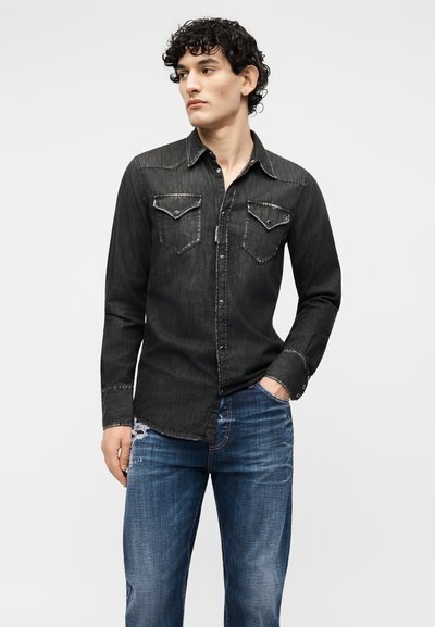 Jeune homme aux cheveux bouclés portant une chemise en jean noire et un jean bleu déchiré, debout avec une main dans la poche, regardant vers la gauche.