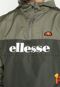 Olivově zelená bunda s kapucí vyrobená z lehkého materiálu, s předním zipem a bílým logem "ellesse" přes hruď, doplněná o malý oranžový akcent.