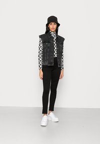 Colete de ganga preto sobre uma camisa de padrão geométrico preto e branco, combinado com leggings pretas e sapatilhas de plataforma brancas. Chapéu bucket preto.