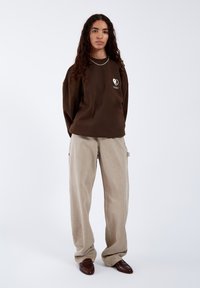 Felpa oversized marrone con un piccolo logo a forma di cuore bianco, abbinata a pantaloni beige larghi e scarpe marroni texturizzate. Design semplice e tonalità neutre.