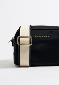 Sort satin taske med en cremfarget vævet rem og guldfarvet hardware. Den har en lynlåslukning og branding i guld på forsiden.