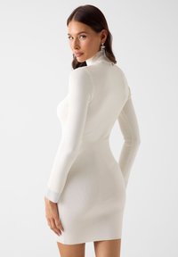 Robe en tricot blanche à silhouette ajustée, manches longues et col montant. Elle présente une texture côtelée et des poignets décorés de motifs pour plus de détails.