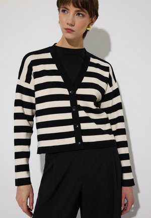 Jeune personne portant un cardigan rayé horizontal noir et blanc par-dessus un haut noir et un pantalon noir, regardant légèrement sur le côté.