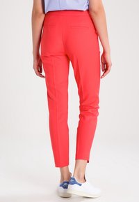 Pantalon rouge slim-fit en tissu lisse, muni de deux poches arrière, avec des jambes fuselées, associé à des baskets blanches avec des accents bleus.