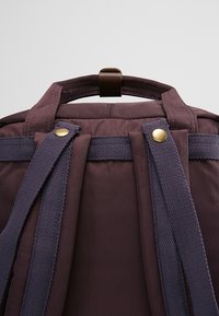 Zbliżenie górnej tylnej części ciemnofioletowego plecaka z materiałowymi paskami na ramiona i małą pętlą z brązowej skóry.