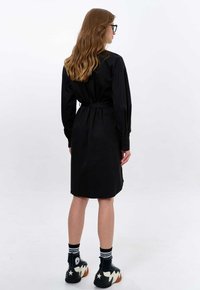 Schwarzes Hemdkleid mit langen Ärmeln, tailliertem Schnitt und Knopfmanschetten. Kombiniert mit schwarzen Sneakern und gestreiften Socken. Glatte Textur.