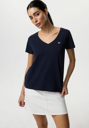 Kobieta z ciemnymi włosami związanymi w kucyk, ubrana w granatowy T-shirt z dekoltem w serek i białą spódnicę, stojąca na tle jednolitego jasnego tła.