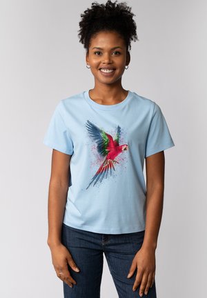 T-shirt en coton bleu clair avec un graphique de perroquet coloré aux accents rouges, verts et bleus. Manches courtes et col rond.