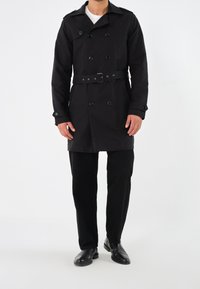 Svart trenchcoat i slät tyg, med dubbelknäppt knappslå, bälte i midjan och axelklaffar. Matchas med svarta byxor och skor.