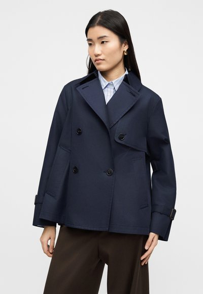 Γυναίκα με μακριά σκοτεινά μαλλιά που φορά χρυσούς κρίκους στα αυτιά, μπλε διπλόφτερη ζακέτα, γαλάζια ριγέ πουκάμισο και καφέ παντελόνι με φαρδιά πόδια.