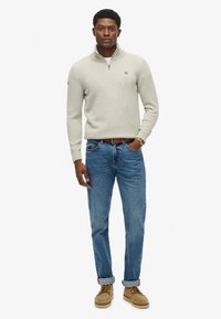 Suéter de punto gris claro con cuello de cremallera, combinado con jeans azules y zapatos color tostado. El suéter cuenta con puños acanalados y un logo en el pecho.