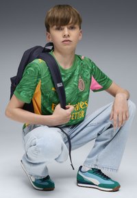 Maglietta sportiva grafica verde con accenti gialli e rosa, abbinata a jeans di jeans blu chiaro e sneaker turchesi. Zaino nero indossato su una spalla.