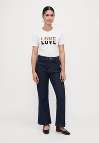 Kvinna som bär en vit t-shirt med texten "LOVE", mörkblå jeans med framfickor och svarta låga klackskor, stående mot en enfärgad bakgrund.
