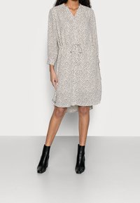 Kurzärmeliges, knielanges Kleid aus leichtem Stoff mit schwarzem Punkte-Muster. Verfügt über eine taillierte Schnittführung und einen V-Ausschnitt, kombiniert mit schwarzen Knöchelstiefeln.