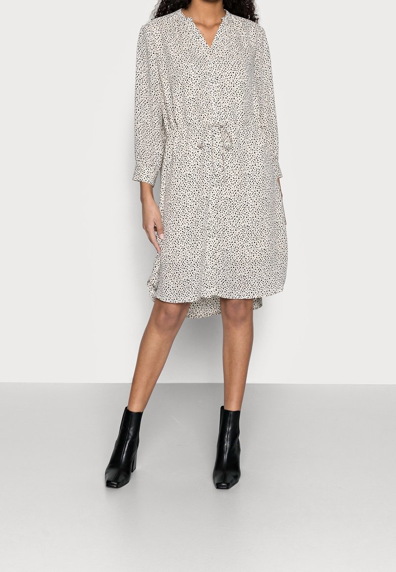 Kurzärmeliges, knielanges Kleid aus leichtem Stoff mit schwarzem Punkte-Muster. Verfügt über eine taillierte Schnittführung und einen V-Ausschnitt, kombiniert mit schwarzen Knöchelstiefeln.