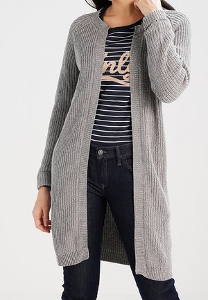 Frau trägt eine lange graue Strickjacke über einem schwarz-weiß gestreiften Hemd mit beigem Text, kombiniert mit dunkelblauen Jeans.