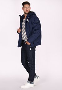 Giacca impermeabile blu navy con cappuccio, cordini bianchi, camicia a righe, jeans in denim scuro e sneaker bianche. Design semplice e funzionale.