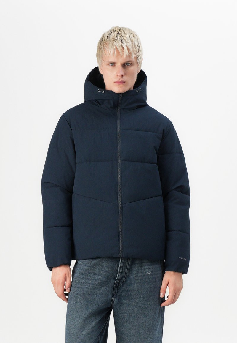 Navy puffer jacket with a hood, featuring horizontal quilted sections, front zipper, and elastic cuffs. Worn with faded jeans.