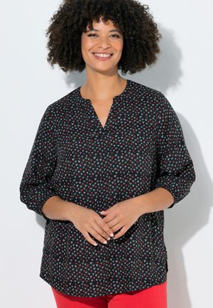 Kvinde med krøllet hår, der smiler, iført en sort bluse med små røde og hvide hjerteeffekter og røde bukser, står op ad en hvid væg.