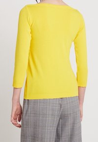 Gul stickad tröja med bred halsringning och trekvartsärmar, ihop med grå rutiga byxor som har en slät yta.