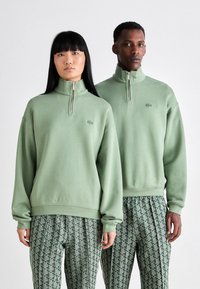 Green, zip-up pullover in a soft material. Ribbed cuffs and hem, featuring a small logo. Modelled with patterned green trousers.