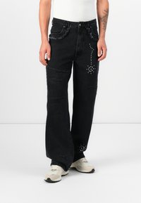Jeans negros desgastados con bordes deshilachados, con acentos de tachuelas plateadas en la pierna derecha. Diseño holgado de cintura alta.