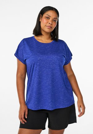 Frau mit schulterlangem schwarzem Haar, die ein blaues kurzärmeliges Hemd und schwarze Shorts vor einem schlichten weißen Hintergrund trägt.