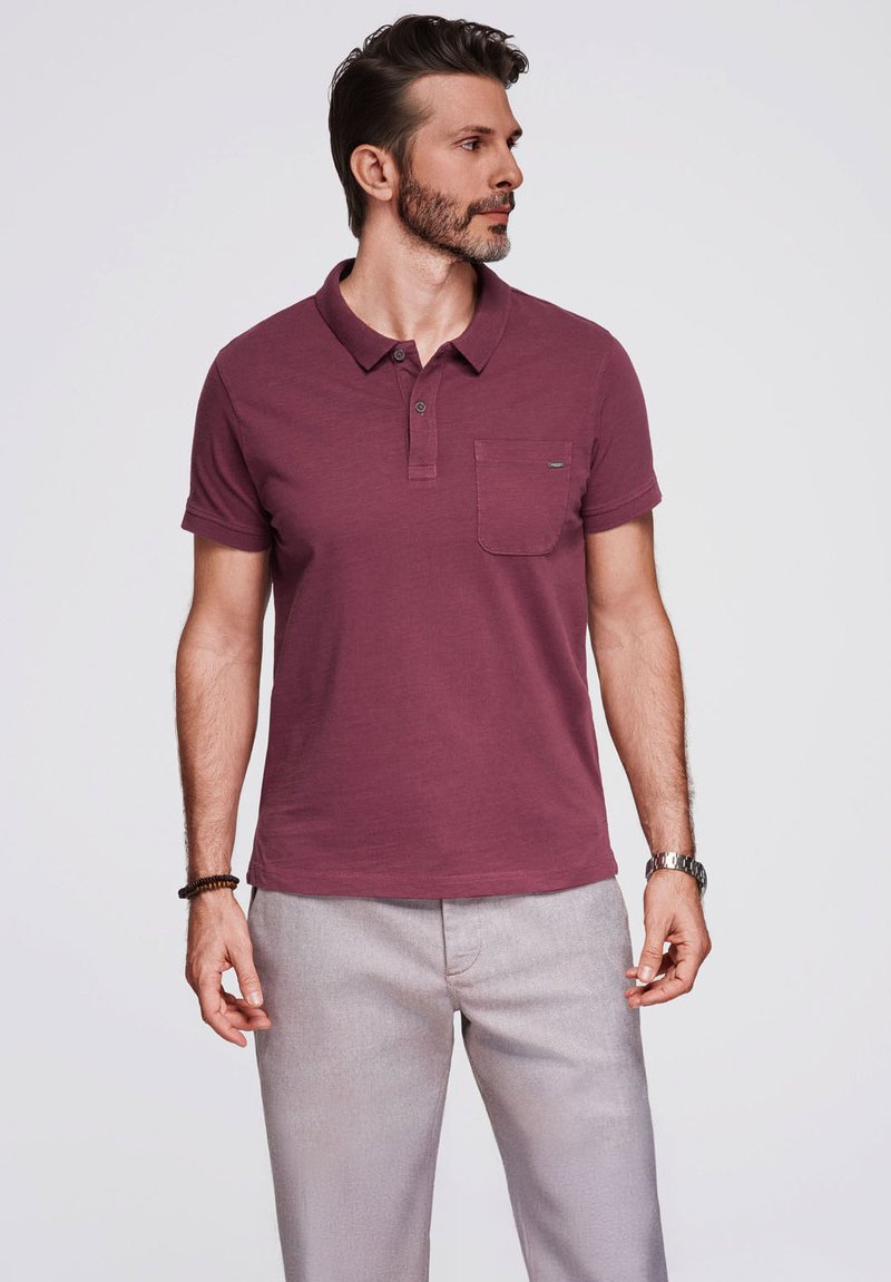 Polo shirt bordeaux con colletto, maniche corte e taschino sul petto; abbinato a pantaloni grigi chiari; texture liscia e design aderente.