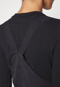 Svart atletisk hettegenser med teksturert stoff, med racerback-design og forsterkede stropper. Stroppene har preget merkevare langs kantene.