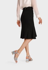 Gonna nera a campana con dettagli cucito, abbinata a sandali con tacco argentati caratterizzati da decorativi intagli floreali e cinturino alla caviglia.