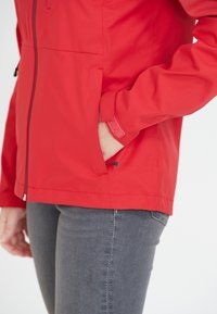 Veste imperméable rouge avec fermeture éclair à l'avant, poches latérales et poignets ajustables. Texture lisse, tissu léger et cordon élastique à l'ourlet.