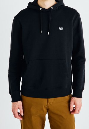 Man draagt een zwarte Lee-hoodie met voorzak en bruine broek, staand tegen een effen witte achtergrond.