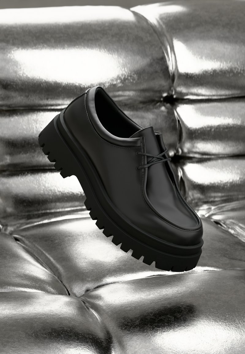 Schwarzer Schnürschuh aus Leder mit dicker, gummierter, profillierter Sohle, präsentiert vor einer glänzenden Metalloberfläche. Glatte Oberfläche, minimalistisches Design.
