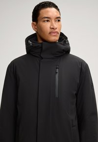 Czarna wodoodporna kurtka z wysokim kołnierzem, regulowanym kapturem i zamkniętą kieszenią na piersi o gładkiej fakturze.