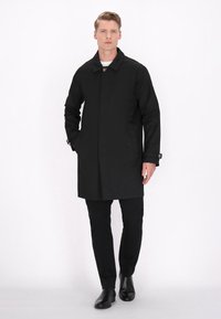 Cappotto lungo nero con colletto abbottonato, tasche laterali e maniche lunghe, abbinato a pantaloni neri e stivali. Tessuto liscio e vestibilità su misura.