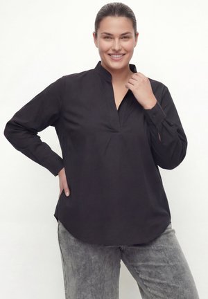 Smiling woman with dark hair tied back wearing a black long-sleeve shirt and gray pants, posing with hand on hip against white background.